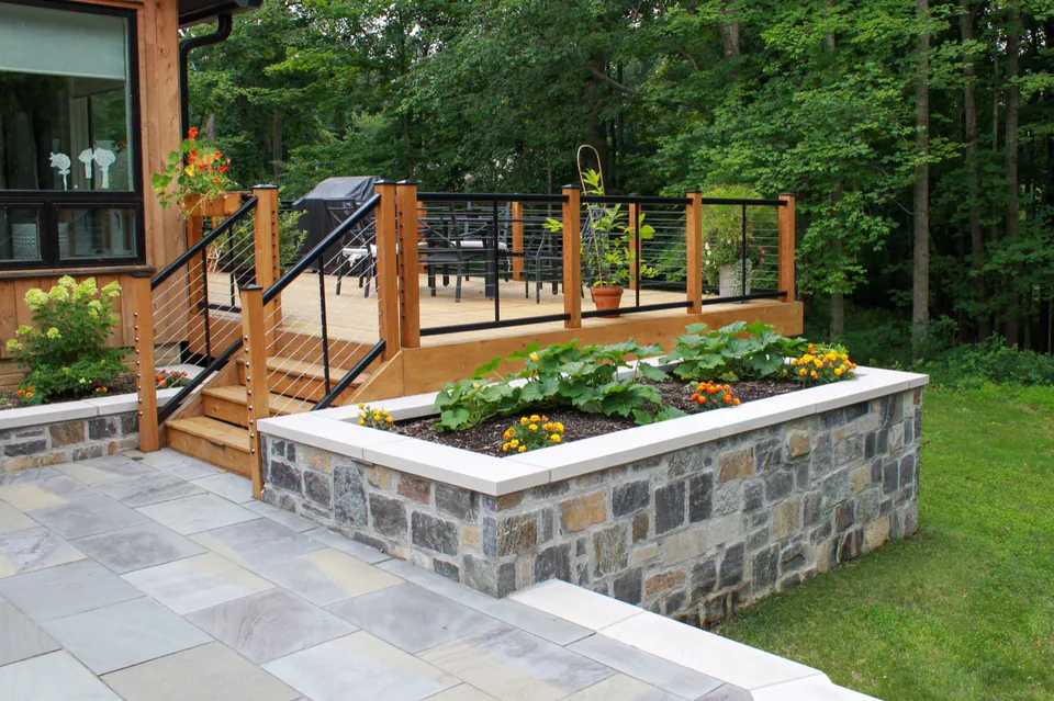 Wood deck stairs leading to stone patio with raised planting bed, stone wall, cable rail, and wooded lawn.